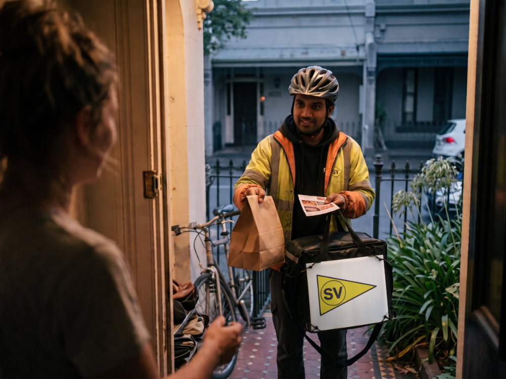 Delivery rider handing food and a flyer to a customer at her front door, StreetValue panel on the bag